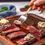 A close-up of perfectly cooked, medium-rare steak slices served on a wooden cutting board. The steak is topped with a dollop of garlic butter and garnished with fresh herbs. A hand holding a fork is lifting a bite-sized piece of steak.
