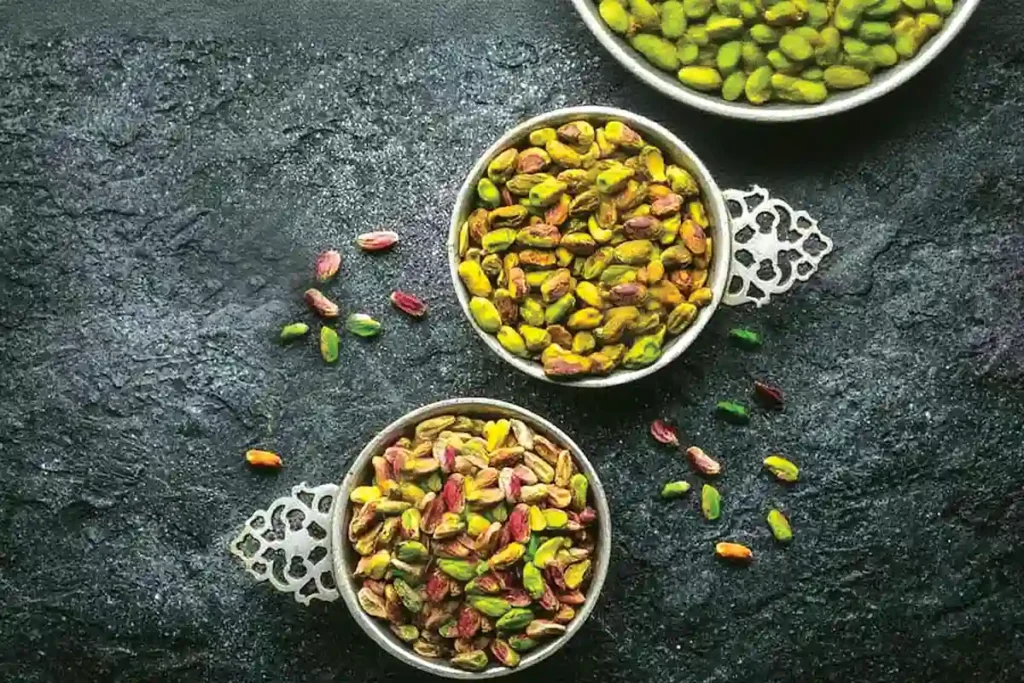 Three bowls of raw pistachios in shells, arranged on a dark textured surface with scattered nuts around them.