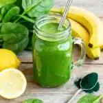 Green detox smoothie in a mason jar with fresh spinach, banana, lemon, and spirulina powder on a rustic wooden table.