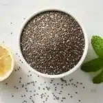 Benefícios da chia: tigela branca com sementes de chia, limão cortado e folhas de hortelã fresca sobre mármore branco.