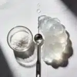 Como preparar gelatina com técnica de blooming: tigela de vidro com gelatina em po seco ao lado de massa de gelatina completamente hidratada e translucida com gotas de agua e colher dosadora sobre marmore branco