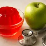 Truque da gelatina para emagrecer: gelatina vermelha sem açúcar em tigela de vidro ao lado de maçã verde e estetoscópio sobre mármore branco.