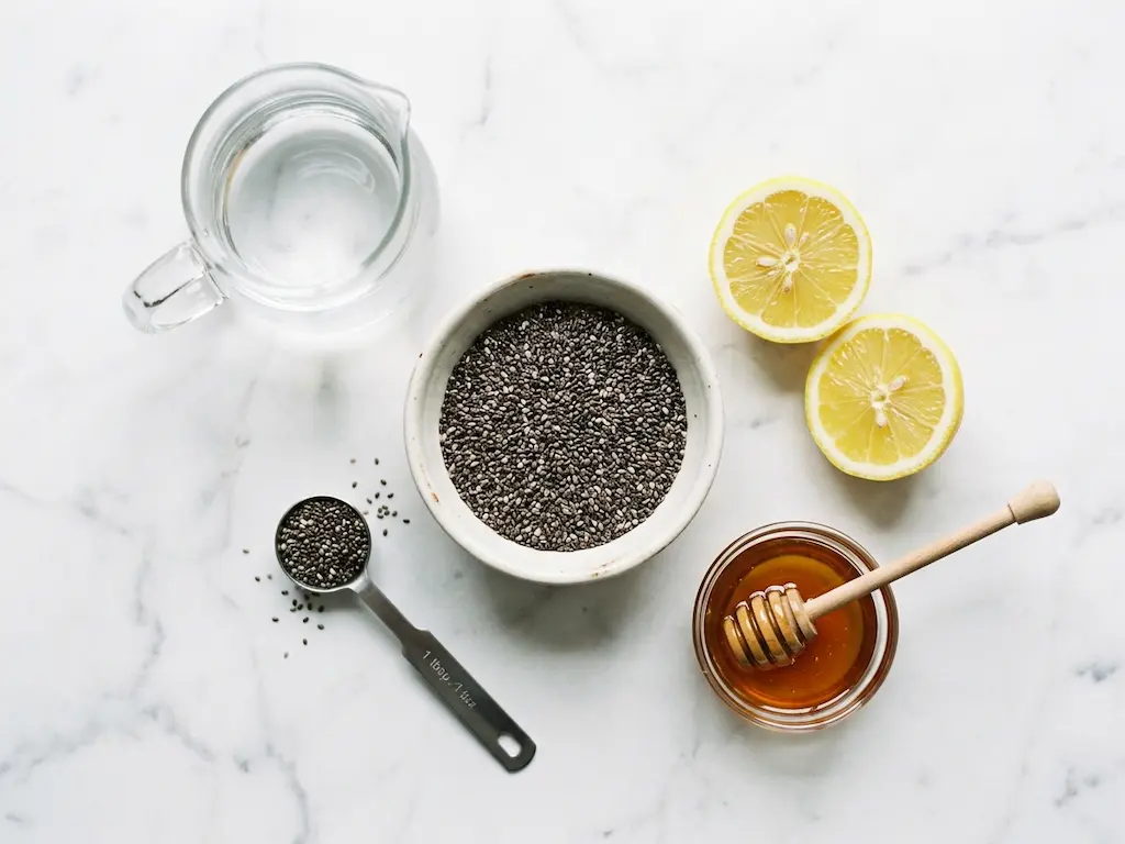 Chia seed water for weight loss ingredients flat lay showing a bowl of chia seeds, a measuring spoon, fresh lemon, honey, and a pitcher of water on white marble.